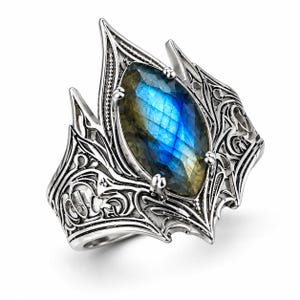 May include: A silver ring featuring a marquise-cut labradorite gemstone with blue and gold iridescence. The band is crafted with intricate, raised metal scrollwork and filigree patterns that extend upward to form a pointed, crown-like setting.
