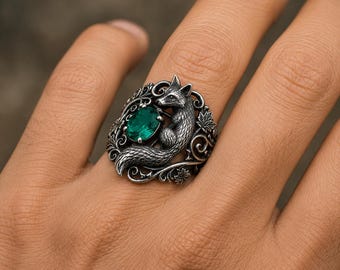 May include: A silver ring featuring a sculpted fox figure integrated into an ornate band with swirling filigree and maple leaf details. An oval-shaped, faceted emerald green gemstone is set securely beside the fox, creating a detailed metalwork design.