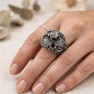 Könnte beinhalten: Ein silberner Ring mit einer Frosch-Darstellung auf Seerosenblättern und einem kleinen roten Edelstein. Der Ring hat detaillierte Muster und einen Vintage-Stil. Der Froschring ist ein einzigartiges Schmuckstück.