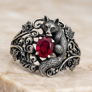 May include: A silver ring featuring a detailed fox figure integrated into a band of ornate scrollwork and maple leaf motifs. The centre of the ring showcases a prominent, oval-cut deep red gemstone held in place by four small metal prongs.