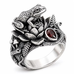 May include: A silver ring featuring a detailed frog perched atop a lotus flower. The band is textured like tree bark and decorated with small mushrooms, fern fronds, and a single teardrop-shaped dark red gemstone set in a silver bezel.