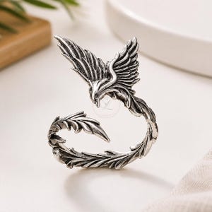 May include: A silver-toned ring featuring a detailed phoenix design. The ring showcases the bird's wings and head, with a curved band adorned with leaf-like details. The ring is set against a white background.