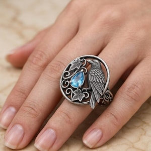 May include: A silver-toned ring featuring a detailed raven perched beside a teardrop-shaped, light blue gemstone. The raven is set within a circular frame with leaf accents. The ring is worn on a finger.