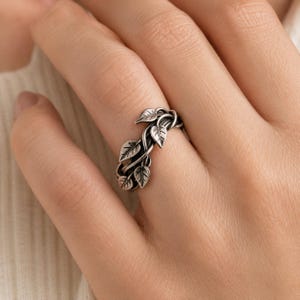 May include: A silver-toned ring featuring a detailed, oxidized metal design of intertwined vines and small leaves that wrap around the band. The metal has a darkened finish in the recessed areas to highlight the intricate leaf textures.