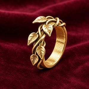 May include: A gold-toned metal ring featuring a textured band designed to resemble intertwined vines. The surface of the ring is adorned with several small, detailed leaf motifs that wrap around the exterior of the band, creating a continuous organic pattern.