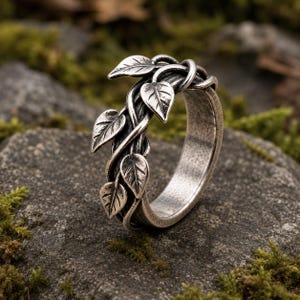 May include: A silver-toned metal ring featuring a band designed to resemble intertwined vines with several small, detailed leaves extending from the surface. The metal has a darkened, oxidized finish in the recessed areas to highlight the texture.