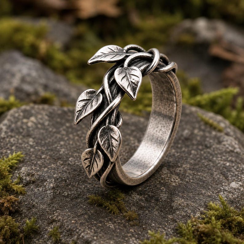 May include: A silver-toned metal ring featuring a band designed to resemble intertwined vines with several small, detailed leaves extending from the surface. The metal has a darkened, oxidized finish in the recessed areas to highlight the texture.