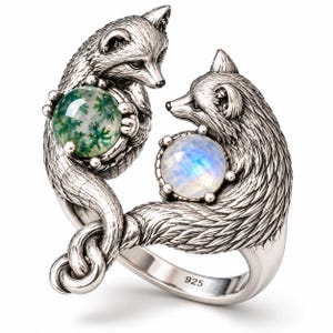 May include: A sterling silver bypass ring featuring two detailed fox figures facing each other. One fox holds a round green moss agate cabochon, while the other holds a round iridescent moonstone. The band is stamped with 925 hallmark.