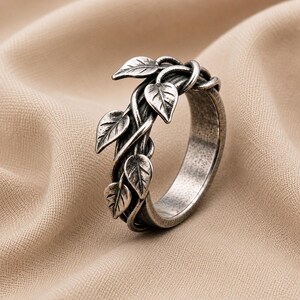 May include: A silver-toned ring featuring a band designed to resemble intertwined vines with multiple small, detailed leaves extending from the surface. The metal has a darkened, oxidized finish that highlights the texture of the leaves and the vine-like structure of the band.