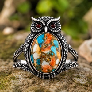 May include: A silver owl ring with a large oval stone. The stone has a mosaic of blue, orange, and white colours. The owl has red gemstone eyes and detailed feathering. The ring's band has decorative scrollwork.
