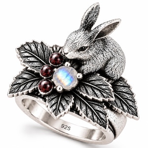 May include: A silver ring featuring a detailed rabbit figure perched atop a cluster of textured leaves. The design includes three small round dark red stones and one central round iridescent moonstone. The inner band is clearly stamped with 925.