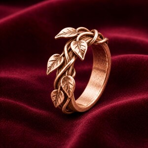 May include: A copper-toned metal ring featuring a band designed to resemble intertwined vines with several small, detailed leaves attached. The surface of the metal has a textured, slightly weathered finish throughout the entire piece.