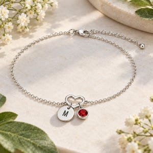 Peut inclure: Un bracelet à chaîne argentée doté d'un petit pendentif en forme de cœur ouvert, d'un disque gravé de la lettre M et d'une pierre précieuse rouge ronde. Le bracelet comprend un fermoir mousqueton et une chaînette d'extension réglable.