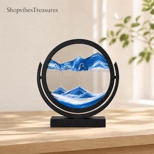 May include: A circular sand art display featuring a black frame and base with the text ShopvibesTreasures visible. The glass center contains layers of blue and white sand forming mountain-like peaks and valleys within the round enclosure.