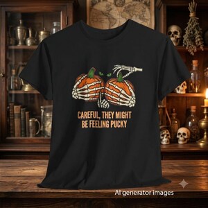 Può includere: Una t-shirt nera con una grafica di due zucche arancioni tenute da mani scheletriche. Sopra le zucche ci sono due occhi verdi luminosi. Il testo sottostante recita CAREFUL, THEY MIGHT BE FEELING PUCKY in un carattere arancione chiaro.