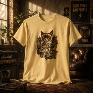 May include: A light yellow t-shirt featuring a graphic of a grumpy cat surrounded by roses and a banner that reads "I'm not anti-social, I am intolerant of shitty people." The shirt is displayed on a patterned rug.