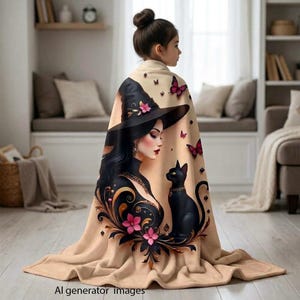 Può includere: Una coperta in pile beige con una stampa grafica di una persona che indossa un cappello da strega nero e un gatto nero. Il design include fiori rosa, elementi decorativi neri a spirale e piccole farfalle rosa sparse sul tessuto.
