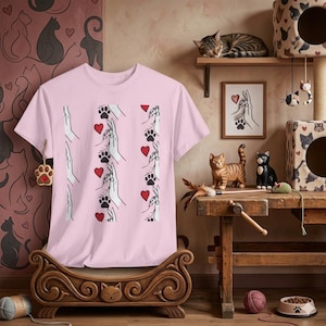 Può includere: Una maglietta rosa chiaro con un motivo ripetuto di mani, impronte di zampe e cuori rossi. Il design è in bianco e nero, con i cuori che forniscono un tocco di colore. La maglietta è esposta su un letto decorativo in legno a tema gatto.