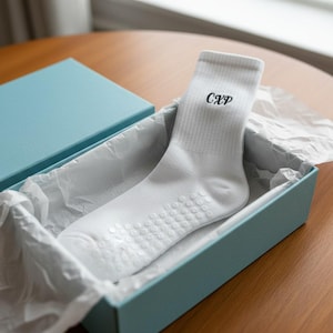 May include: A white crew sock with the black embroidered letters CXP on the ribbed cuff and a pattern of small white circular silicone grips on the sole, presented inside a light blue gift box lined with white tissue paper.