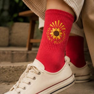 May include: A pair of bright red crew socks featuring an embroidered yellow sunflower graphic centered on the ankle. The text "RADIANT BLOSSOMS" is stitched in a circular pattern around the sunflower in yellow thread. The socks are worn with white canvas sneakers.