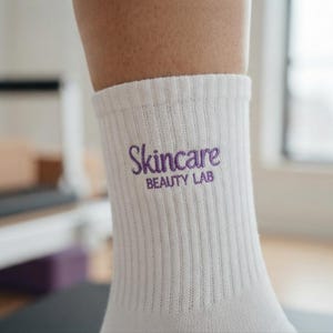 Op de afbeelding: Een witte geribbelde sok met de paars geborduurde tekst Skincare BEAUTY LAB aan de zijkant. De stof heeft een verticale textuur en het ontwerp bevindt zich op het bovenste gedeelte van de sok, die aan het onderbeen wordt gedragen.