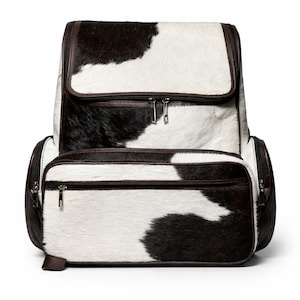 Puede incluir: Una mochila de piel de vaca con un estampado de manchas blancas y negras. La bolsa tiene múltiples compartimentos con cremallera, ribetes de cuero marrón y una solapa superior. La mochila tiene forma rectangular.