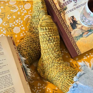 Peut inclure: Paire de chaussettes tricotées à la main, jaunes et vertes. Elles reposent sur un tissu à motifs floraux jaunes, à côté d'un livre intitulé "Anne of Green Gables" et d'une tasse. Un article confortable et fait à la main.