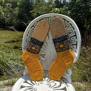 Peut inclure: Une paire de chaussettes tricot&eacute;es jaune et bleu marine avec un motif floral, pr&eacute;sent&eacute;es sur des formes de chaussettes en bois. Les chaussettes sont pos&eacute;es sur une chaise blanche orn&eacute;e &agrave; l'ext&eacute;rieur.
