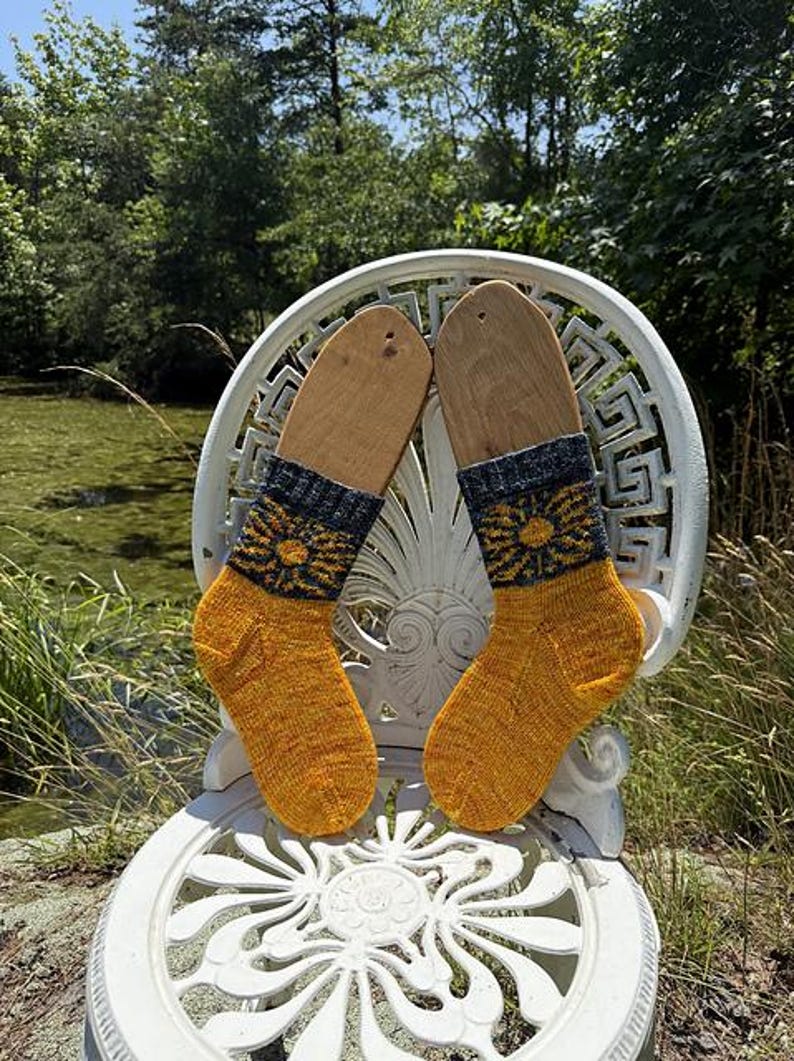 Peut inclure: Une paire de chaussettes tricot&eacute;es jaune et bleu marine avec un motif floral, pr&eacute;sent&eacute;es sur des formes de chaussettes en bois. Les chaussettes sont pos&eacute;es sur une chaise blanche orn&eacute;e &agrave; l'ext&eacute;rieur.