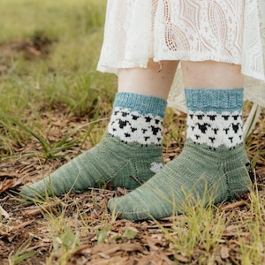 Pode incluir: Um par de meias de tornozelo com a parte do pé em verde, uma faixa branca com padrão de ovelhas pretas e um punho canelado azul. As meias apresentam uma textura de malha visível e uma pequena etiqueta preta na lateral do calcanhar.