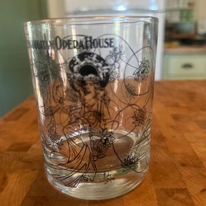 May include: A clear glass tumbler featuring a black line art illustration of a figure surrounded by lotus flowers and circular patterns. The text "METROPOLITAN OPERA HOUSE" is printed near the top rim, and a small signature is visible at the base.