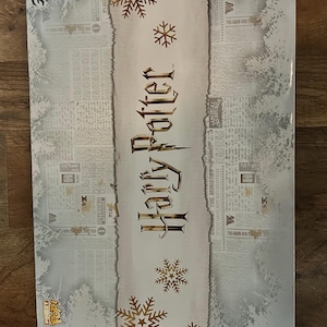 May include: A rectangular box featuring the words "Harry Potter" in gold lettering on a cream-colored banner. The box has a newspaper-style background with gold snowflake accents and the Funko Pop! logo.