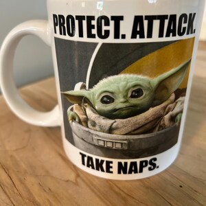 May include: A white ceramic mug featuring a graphic of a green, large-eared creature sitting in a metallic pod. The text "PROTECT. ATTACK." is printed in black above the image, and the text "TAKE NAPS." is printed in black below the image.