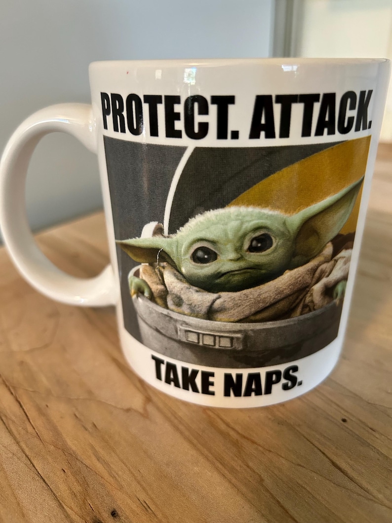 May include: A white ceramic mug featuring a graphic of a green, large-eared creature sitting in a metallic pod. The text "PROTECT. ATTACK." is printed in black above the image, and the text "TAKE NAPS." is printed in black below the image.