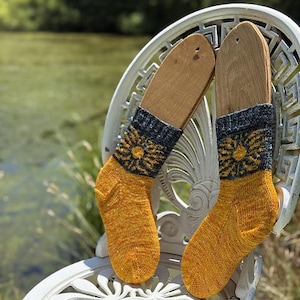 May include: A pair of hand-knitted socks displayed on wooden forms, resting on a white decorative chair. The socks feature a yellow and orange body with a dark blue cuff and a yellow floral pattern. The background shows a blurred view of a pond.