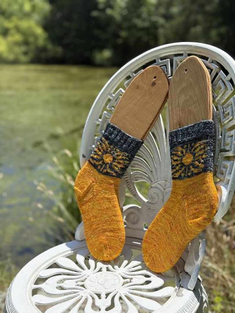 Peut inclure: Une paire de chaussettes tricot&eacute;es &agrave; la main, pr&eacute;sent&eacute;es sur des formes en bois, reposant sur une chaise d&eacute;corative blanche. Les chaussettes pr&eacute;sentent un corps jaune et orange avec un revers bleu fonc&eacute; et un motif floral jaune. L'arri&egrave;re-plan montre un &eacute;tang flou.