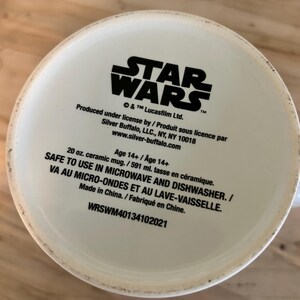 May include: The bottom of a white ceramic mug featuring black printed text that reads STAR WARS, Produced under license by Silver Buffalo, LLC., NY, NY 10018, www.silver-buffalo.com, Age 14+, 20 oz. ceramic mug, SAFE TO USE IN MICROWAVE AND DISHWASHER.