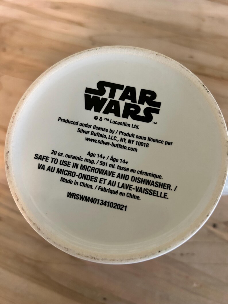 May include: The bottom of a white ceramic mug featuring black printed text that reads STAR WARS, Produced under license by Silver Buffalo, LLC., NY, NY 10018, www.silver-buffalo.com, Age 14+, 20 oz. ceramic mug, SAFE TO USE IN MICROWAVE AND DISHWASHER.