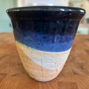 May include: A small ceramic cup featuring a two-tone design with a glossy dark blue glaze on the upper portion and a textured, speckled beige and light orange finish on the lower section, separated by a distinct wavy line around the body.