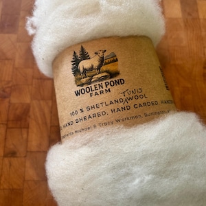 May include: A ball of fluffy white wool wrapped in a tan paper label. The label features a graphic of a sheep and trees with the text Woolen Pond Farm, 100% Shetland Wool, Tunis, Hand Sheared, Hand Carded, Hands, Shepherds Michael & Tracy Workman.
