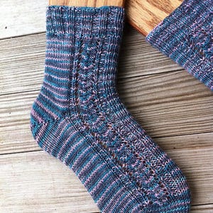 Peut inclure: Une chaussette tricotée à rayures bleues, roses et grises. La chaussette présente un motif ajouré décoratif sur le devant et est fabriquée en fil doux. Le bord-côte est côtelé et la chaussette est présentée sur une surface en bois.