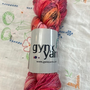 May include: A skein of hand-dyed yarn in shades of pink, red, orange, and purple. The yarn is wrapped with a label that reads "gynx yarn" and the website address. The yarn is displayed on a white embroidered cloth.