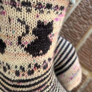 May include: Close-up of a knitted sock with a cream-colored base, featuring black cat silhouettes and dark grey floral patterns. The sock has black and white striped sections and flecks of pink and blue.