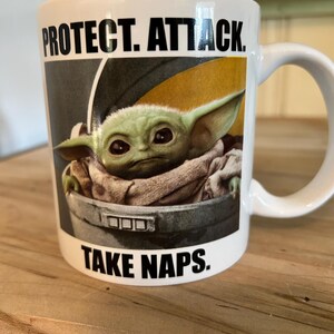 May include: A white ceramic mug featuring a graphic of a green creature with large ears sitting in a gray pod. The text above the image reads PROTECT. ATTACK. and the text below the image reads TAKE NAPS. The mug has a curved handle on the right side.