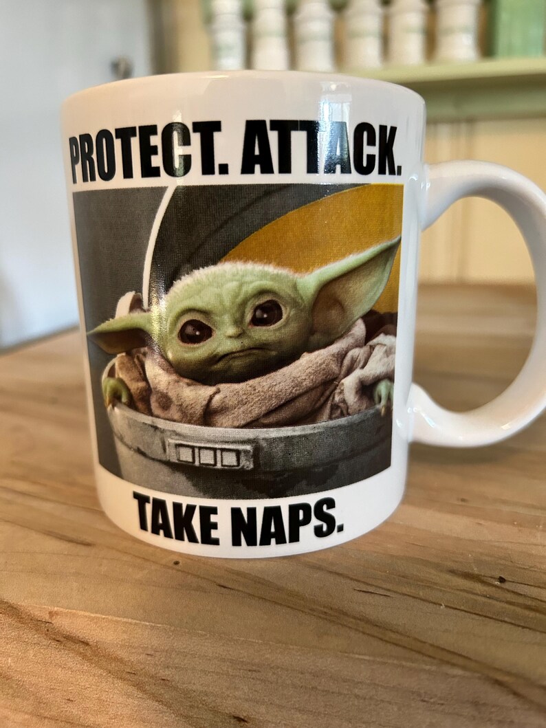 May include: A white ceramic mug featuring a graphic of a green creature with large ears sitting in a gray pod. The text above the image reads PROTECT. ATTACK. and the text below the image reads TAKE NAPS. The mug has a curved handle on the right side.