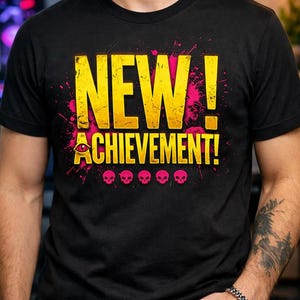 May include: A black short-sleeved t-shirt featuring the yellow text NEW! ACHIEVEMENT! with a pink eye icon replacing the letter A. The design includes pink paint splatter effects and a row of five small pink skull icons centered below the text.