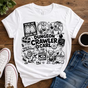 May include: A white t-shirt featuring a black graphic print with the text Dungeon Crawler Carl. The design includes illustrations of a dinosaur, a goat, a cat riding a dinosaur, a goblin, a treasure chest, a skull, a donut, and various symbols.