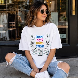 Peut inclure: T-shirt blanc avec un motif coloré de tuiles de Mahjong et le texte "CRAKS DOTS BAMS IYKYK". Le t-shirt est porté avec un jean bleu. La personne porte des lunettes de soleil.