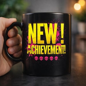Może przedstawiać: Czarny ceramiczny kubek z żółtym napisem NEW! ACHIEVEMENT!, w którym stylizowana ikona oka zastępuje literę A. Projekt zawiera różowe plamy farby oraz rząd pięciu małych różowych ikon czaszek w dolnej części grafiki.