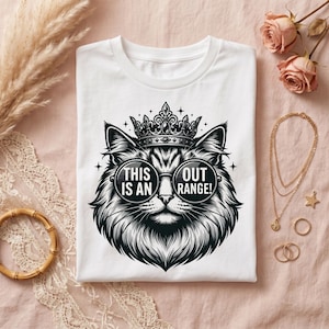 May include: A white t-shirt featuring a black and white graphic of a cat wearing a crown and round sunglasses. The text on the lenses reads "THIS IS AN" on the left and "OUT RANGE!" on the right. The design is centered on the front of the shirt.